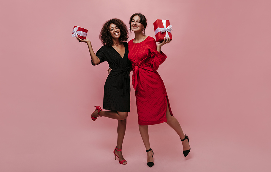 Two charming fashionable girls with brunette hair in stylish polka dot red and black dresses and heels holding gift boxes, smiling and hugging.